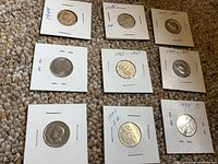 Nine coins displayed on a carpet, each in their own white coin holder with dates and other information handwritten on holders.