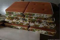 Stack of 8 vintage patio cushions showing burnt orange tops with button tufting and floral sides with white piping.