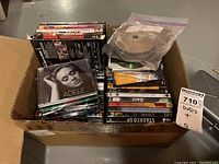 Photo of a large cardboard box filled with many DVDs and some music CDs. DVDs mostly in cases with visible titles, some discs loose in plastic bags. CDs include Adele 21 and others.