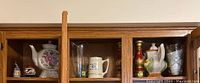 Wooden shelf containing various glass and ceramic serving vessels including a teapot, mug, glass pitcher, and decorative bottles.
