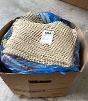 Cardboard box filled with items for sale, topped with a woven beige netted mat and some blue wrapping inside the box.