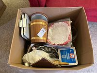 Cardboard box filled with various kitchen items including fabric oven mitt, metal trays, plastic container, and vintage carving set.