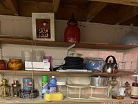 Three shelves containing various housewares including a red ceramic jar, metal utensils, cleaning products, and decorative tins.