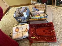 Photo showing boxes filled with assorted linens including tablecloths, throws, and bedding fabrics with varied patterns, next to a small decorative rug.