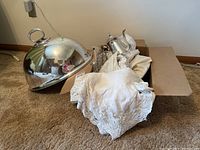 Silver tone metal dome food cover with handle, silver teapot and matching accessories partially wrapped in lace linens inside a cardboard box on carpet.