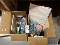 Wide view of two open cardboard boxes with various small items inside, including rosaries, white binder, and other household items.