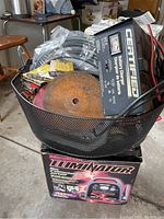 Mesh basket containing assorted tools, sockets, screwdriver bits, staples, a rusty circular tool disc, and wires on top of MotorMax Eliminator power pack box.