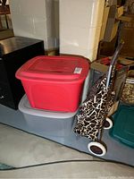 Red Tuff brand storage box stacked on gray plastic tote next to a giraffe print rolling travel bag buggy with a metal frame.