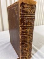 Image of the Bible standing upright showing dark brown leather cover with gold embossed decorations and text 'Holy Bible' on spine, signs of edge wear.