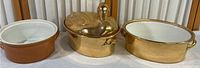 Three Hall crocks displayed side by side: brown glazed ground crock, golden plated duck crock with detailed duck head handle, and golden plated round crock with two side handles.