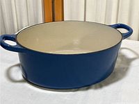 Front angle view showing full body and handles of blue oval Dutch oven with cream interior on white cloth background.