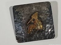 Front view of the sterling silver square brooch with hammered texture and gold-tone flute player applique centered on front.