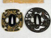 Two samurai sword hilts, one rectangular with floral gold design and one round with dragon design, laid flat on white surface.