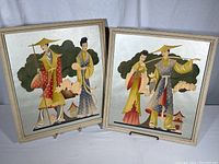 Two framed paintings showing Japanese couples in traditional dress against silver background, both standing on easels.