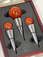 Three wine bottle stoppers with polished metal tapered bodies and round wood ball tops in a foam insert inside a wood box.