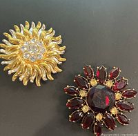 Two gold-tone flower shaped vintage brooches, one with clear rhinestones in sunburst pattern, one with dark red faceted stones in floral pattern