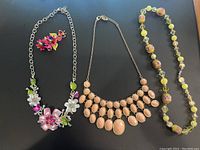 Photo shows complete lot of 3 necklaces and 1 brooch arranged flat on black background, detailing style and colors of each piece.