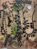 Top-down view of assorted vintage jewelry on cardboard, showing necklaces, earrings, a watch, and bracelets in black, green, gold, and pink tones.