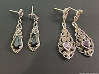 Photo shows two pairs of 925 silver earrings side by side upon black background. Left pair has elongated black faceted stones, right pair has silver openwork with heart-shaped purple stones.