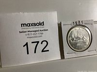 Photo of the coin in a white cardboard holder with MaxSold auction number 172 shown next to it to identify the lot. Shows the front side of the coin with canoe and voyageurs, and year 1965 noted on the holder.