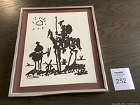 Framed art print showing full artwork and frame, placed on a dark wood surface, with auction lot number visible beside it.