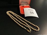 Full view of the pearl necklace arranged in a single strand beside a red velvet box and auction lot number card.