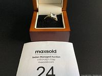 Ring displayed in a wooden box with black velvet insert. Large rectangular clear stone with two small round stones on sides on a gold-tone band visible.