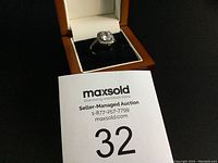 Ring with large cushion-cut center stone and halo setting on metal band, displayed in jewelry box with black velvet interior and tan/brown exterior.