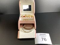 Wide shot of pink jewelry box open showing gold tone 'MOM' pins in upper section and silver bracelet partially visible in lower drawer.