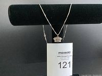 Silver necklace with a crown pendant hanging on a black stand with a tag showing lot number 121.