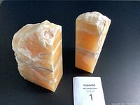 Two onyx stone sculptures showing natural banded layers in shades of orange and white, photographed from front with polished and rough surfaces visible.