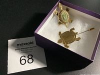 Two turtle brooches displayed in a purple box with white cushion: one ornate gold-tone turtle with green textured stone shell, one simpler gold-tone turtle stick pin brooch.