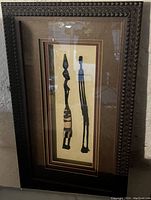 Photo of the 3D framed artwork showing two tall slender African carved figures in a carved dark wood frame with multi-color mats.