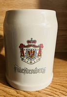 Front view of an off-white ceramic beer stein featuring a colorful Fürstenberg crest and gothic style lettering below it.