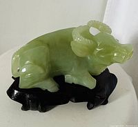 View of a green jade water buffalo sculpture resting on a carved dark wooden stand, showing the full side profile of the buffalo with intricate horns and facial features.