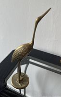 Side angle of brass standing bird with detailed wing etching and round base on mirrored surface.