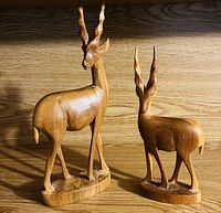 Photo of two handcrafted teak wood antelope figurines on a wooden surface. The larger and smaller figurine display smooth polished surfaces and twisted horn details.