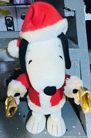 Photo showing plush Snoopy doll in red Santa suit holding gold bells, white feet, and black ears.