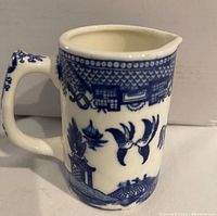 Blue and white porcelain jug with classic Blue Willow pattern depicting birds and oriental buildings, detailed border, and a pouring spout.