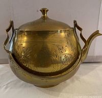 Brass tea pot with floral engravings on bulbous body, side view showing curved spout and handle
