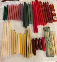 Photo showing a collection of various candles laid out on a white surface. Candles include a variety of colors and styles such as tapered, twisted spiral, pillar, and packaged sets.