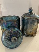 Pair of blue carnival glass candy jars, one hexagonal and one cylindrical with grape and leaf design on body and lids, decorative knob tops.