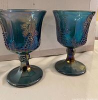 Pair of iridescent blue carnival glass goblets showing grape and leaf embossed pattern, flared rims and decorative stems with grape cluster details.