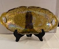 Front view of an oval carnival glass dish on a black stand, showing iridescent gold finish with floral embossing and two handles.