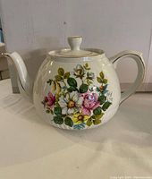 View of the white ceramic tea pot showing the floral print with pink, white, and yellow flowers, and gold trim on handle and lid.