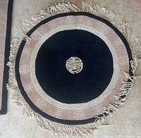 Round floor rug with black center, beige border with Asian motifs, and fringe.