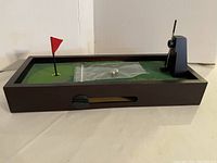 Angled view of the tabletop putting game showing the green felt putting area, red flag, two metal balls in a plastic bag, and the mechanical scoring lever on the side.
