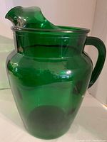 Photo showing a green transparent glass pitcher with a handle and a wide spout, exhibiting a smooth, rounded design.