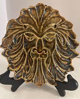 Front view of ceramic lion face trivet with textured layered mane and earthy golden glaze on display stand.