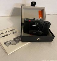 Minox 35 EL camera in open hard case with instruction manual on side, showing front and lens area.